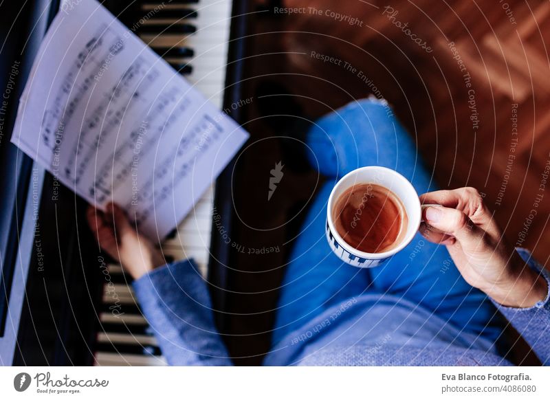 young unrecognizable woman holding a cup of coffee and playing piano by reading a music sheet. Music concept indoors. top view style human key note lesson