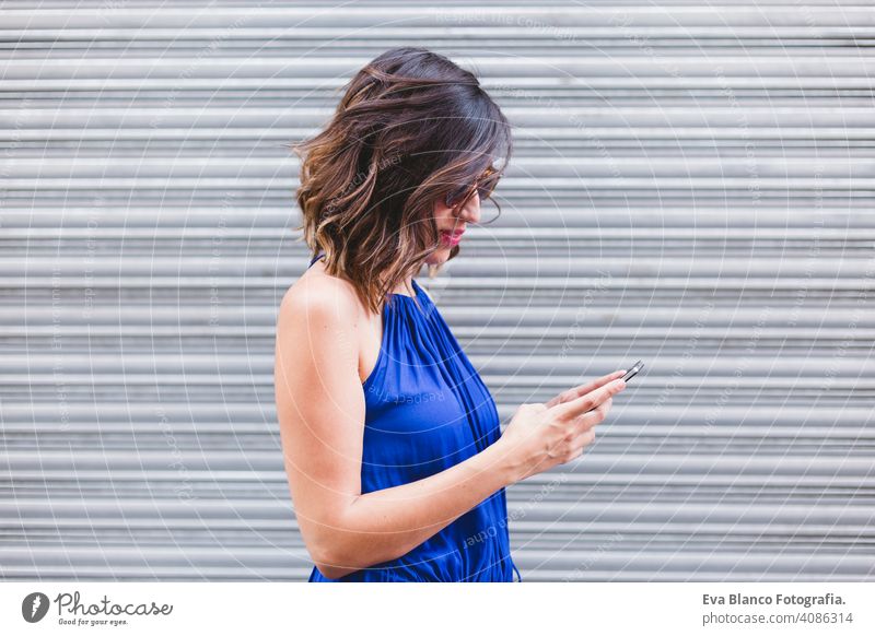 young beautiful woman using mobile phone and smiling. She is in the street, wearing a casual blue dress, sunglasses and smiling. LIfestyle outdoors, modern life and technology