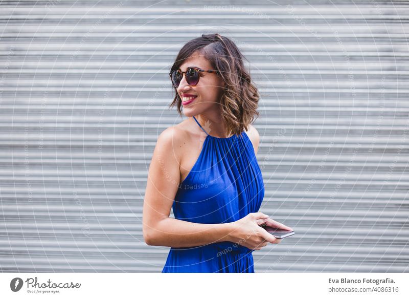 young beautiful woman using mobile phone and smiling. She is in the street, wearing a casual blue dress, sunglasses and smiling. LIfestyle outdoors, modern life and technology