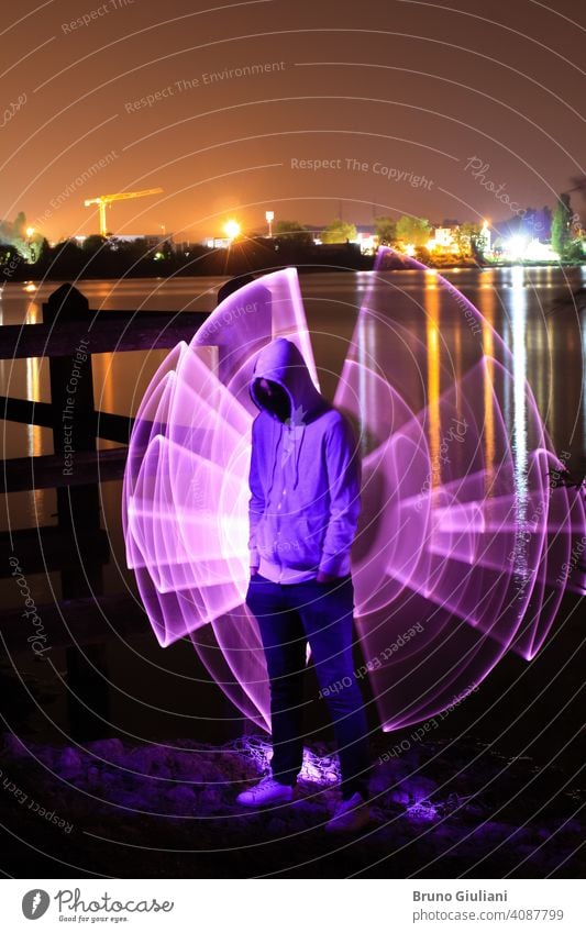 A man standing with his hands in his pockets and a hood over his head, standing at night in front of a lake. Violet abstract shape with light saber behind it. City lights in background.