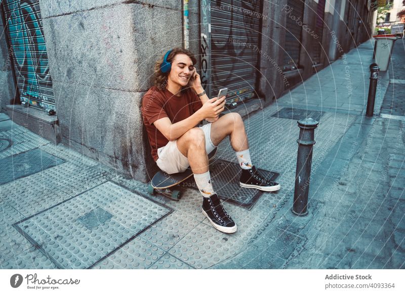 Relaxed man in headphones using smartphone while resting on longboard on street relaxed carefree generation millennial casual browsing sit building city urban