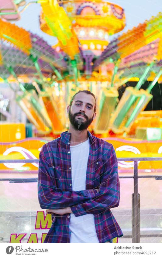 Confident bearded man resting by shiny multicoloured attraction at amusement park confident hipster calm casual serious carousel fairground colourful bright