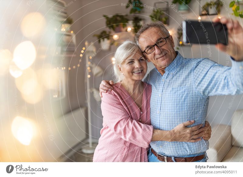 Senior couple in the living room taking selfie with smartphone people woman adult senior mature casual attractive female smiling happy Caucasian toothy enjoying