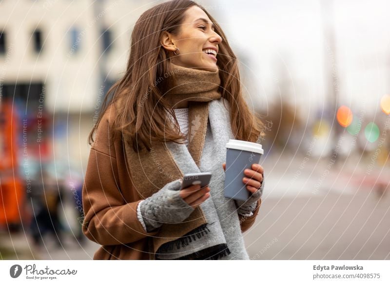 Smiling young woman with smart phone and coffee cup outdoors at urban setting autumn fall girl female beautiful weather coat fashion pretty attractive adult