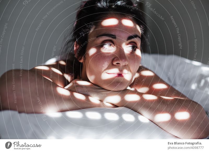portrait of attractive young caucasian woman relaxing in bed during morning time. Lady enjoys fresh soft bedding linen and mattress in bedroom. Blind shadows on face