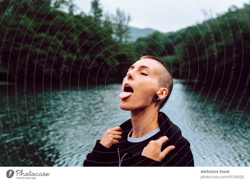 Contemporary woman in casual wear delighting in rain against pond nature enjoy catch raindrop short hair piercing skinhead informal female tongue out green