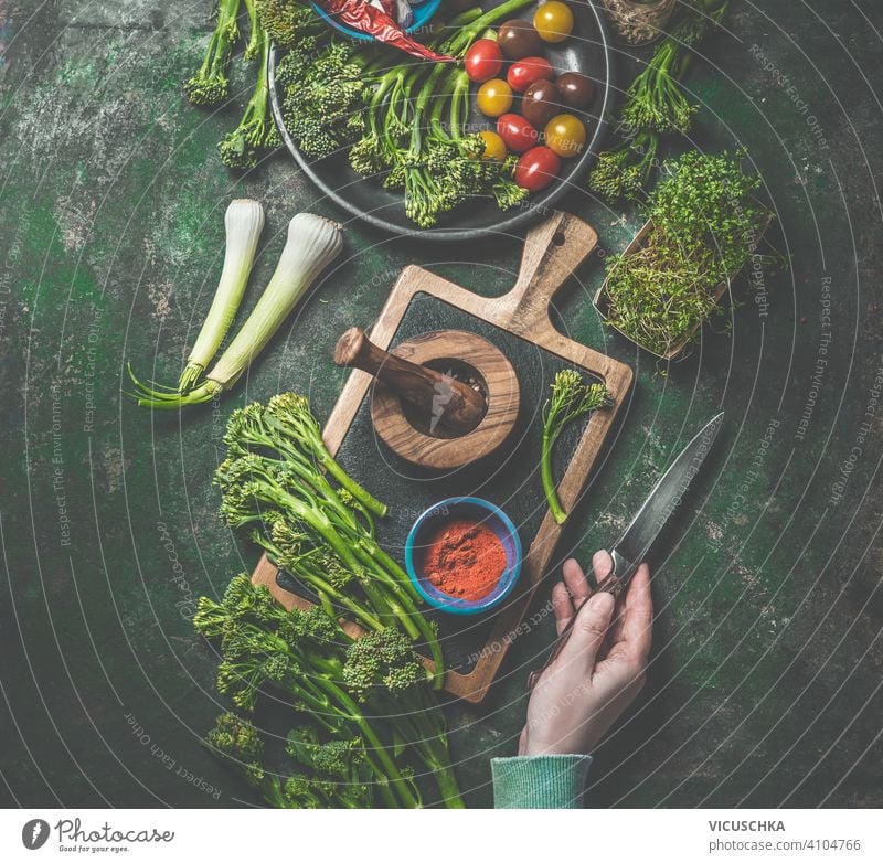 Cooking preparation of wild broccoli. Women hand holding knife on dark rustic background with cutting board and ingredients. Top view. Healthy food cooking