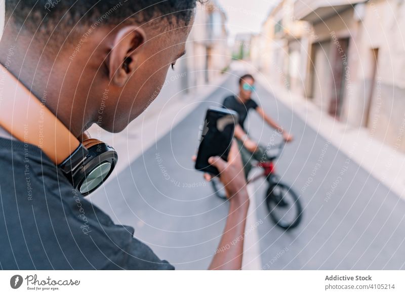 Young black male teenager taking photo on cellphone of friend in street take photo men bmx smartphone cool bike using picture social media communication shot