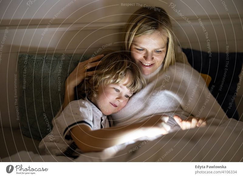 Mother and son using digital tablet together at night in bed bedtime lying down reading bedroom storytelling internet online cute evening sleeping resting boy