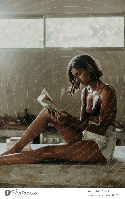 Concentrated young female reading book in kitchen woman enjoy interested comfort pleasure information literature knowledge education domestic hobby bra idyllic