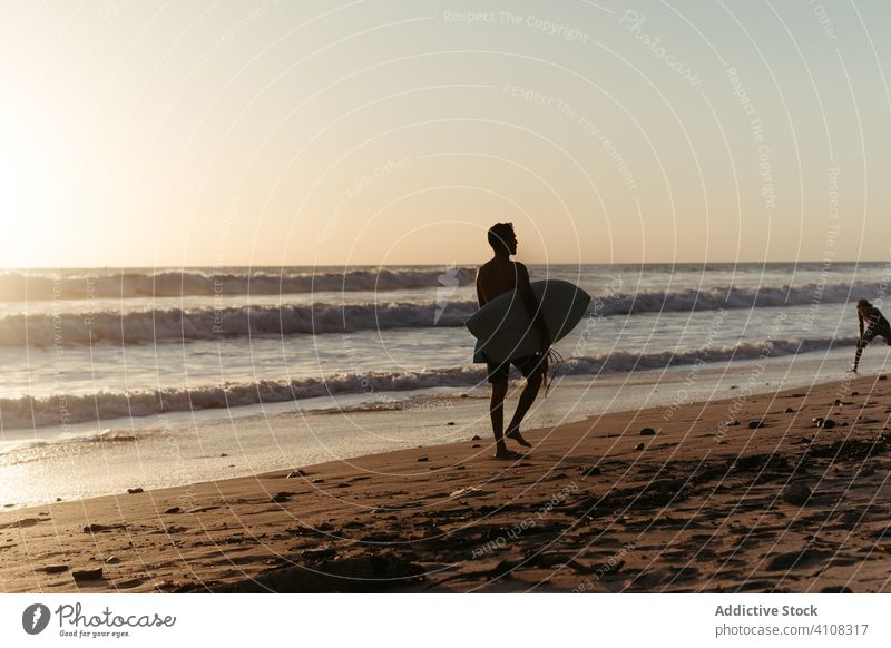 Unrecognizable male surfer walking on beach in sunset man surfboard seashore summertime ocean water surfing wave equipment extreme active sport hobby lifestyle