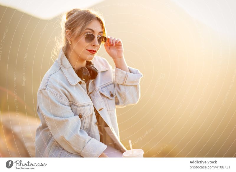 Wistful woman using headphones during relax rest nature sit sunglasses fence casual beverage drink music female style lifestyle holiday young summer journey