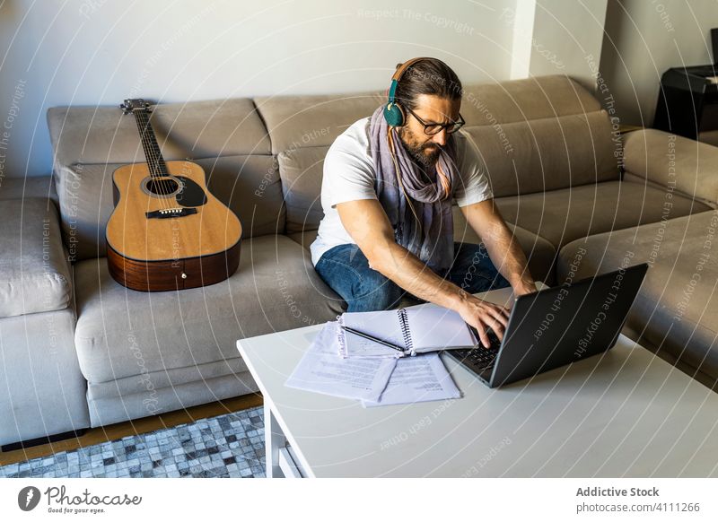 Focused man working with laptop in living room using headphones typing browsing listen songwriter eyeglasses male casual watch connection online poet sit couch