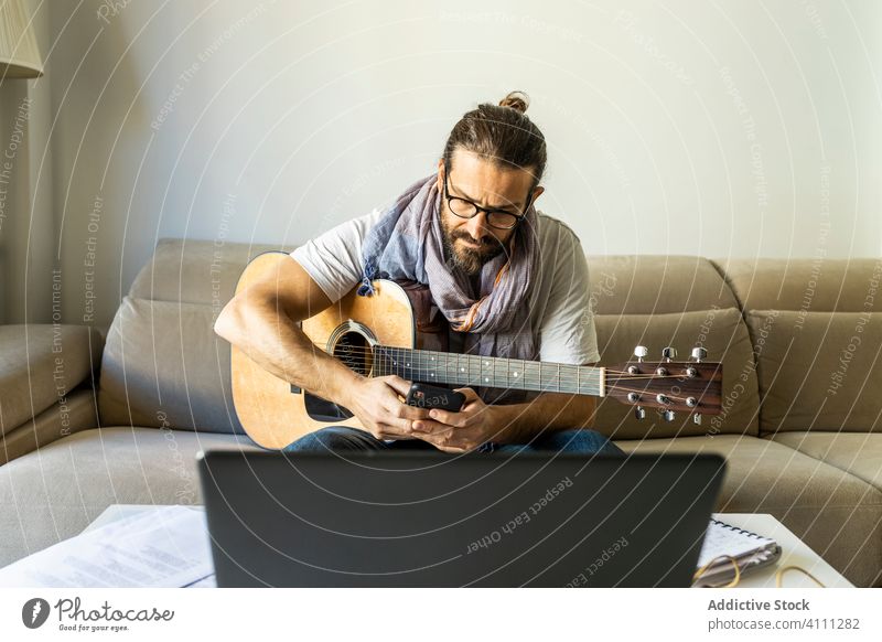 Young male musician browsing smartphone in living room watching talk call guitar using laptop sit couch songwriter composer poet sofa casual communicate man