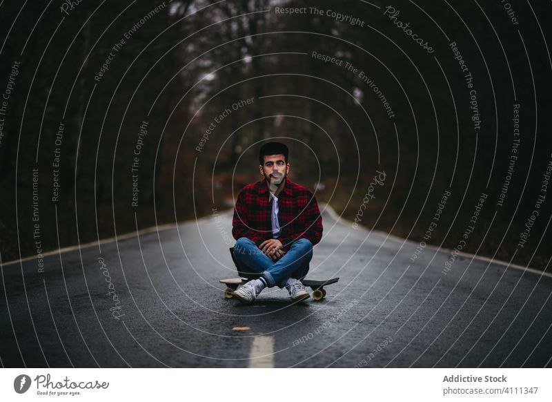 Young man resting on road with skateboard in countryside sit lifestyle male casual young nature asphalt relax forest cool freedom hipster ethnic hobby sport