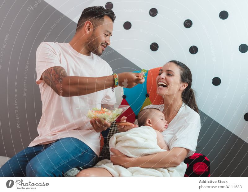 Happy young family with little baby ob armchair mother father sit woman parent love child home care kid talk call speak smartphone feed help mobile eat salad