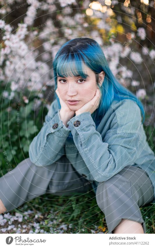 Thoughtful young woman sitting in blooming garden trendy sad teen melancholy dyed hair alternative solitude thoughtful style pensive tree blue hair park flower