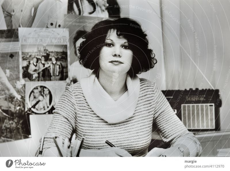 A young woman sits at her desk in the office and looks into the camera. Behind her is an old radio. Artist photos hang on the wall. Woman Young woman