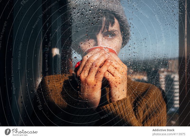 Thoughtful woman drinking coffee near window sad depression self isolation home unhappy lonely covid19 melancholy young female wet rain solitude stress cup