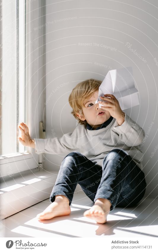 Curious little boy playing with paper plane on white windowsill at home window sill dream curious innocent child barefoot sunny kid childhood happy fun daytime