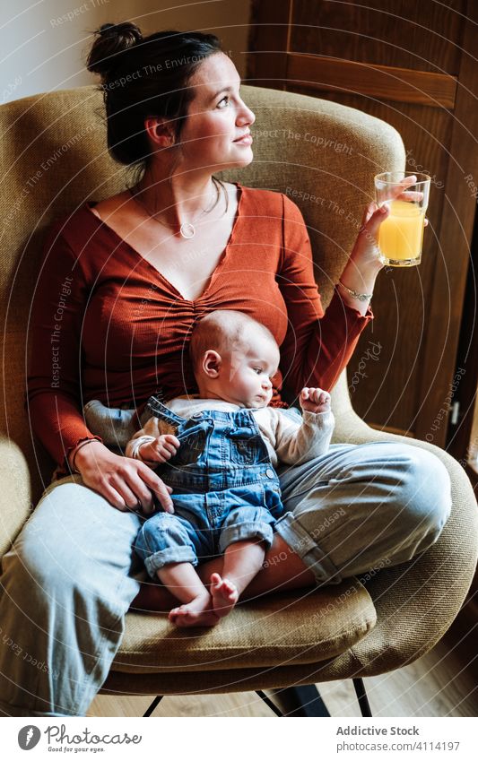 Crop mother with juice and baby resting on chair orange healthy cozy sit love home tender woman fresh drink beverage adorable child kid infant room together