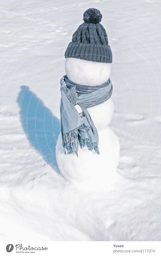 A snowman in a blue hat and scarf is standing in the white field. An iceman on a light showy background. Sunny day, freezing cold, close up. snowy sunny
