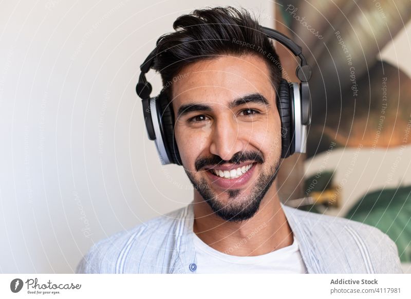 Bearded man listening to music and looking at camera smile home headphones ethnic happy rest weekend male headset cheerful sound beard fun cozy audio cool