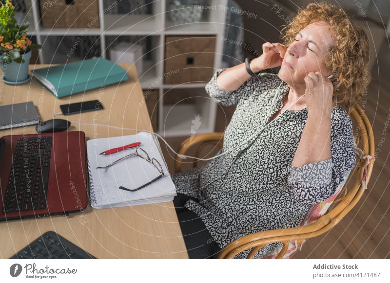 Thoughtful adult woman listening music in earphones while working on laptop at home rest distance remote break enjoy notebook eyes closed using freelance relax