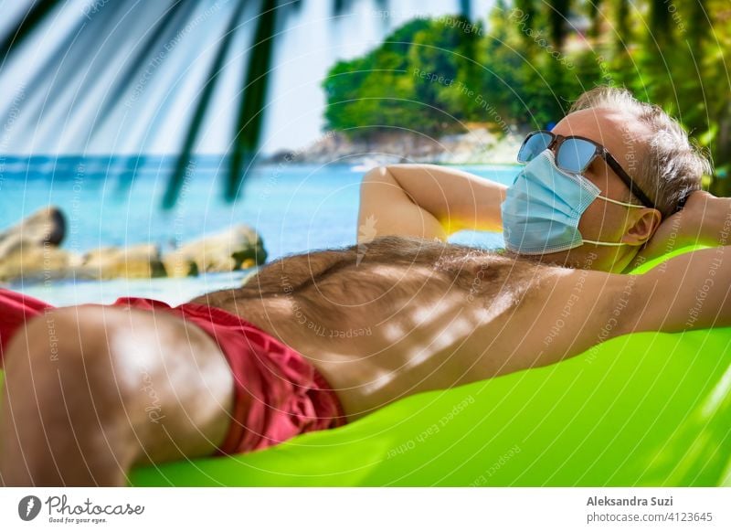 Mature man in protective mask and sunglasses relaxing on beach. Exotic tropic landscape with turquoise water and palms. Tourism during pandemic. Safe traveling.