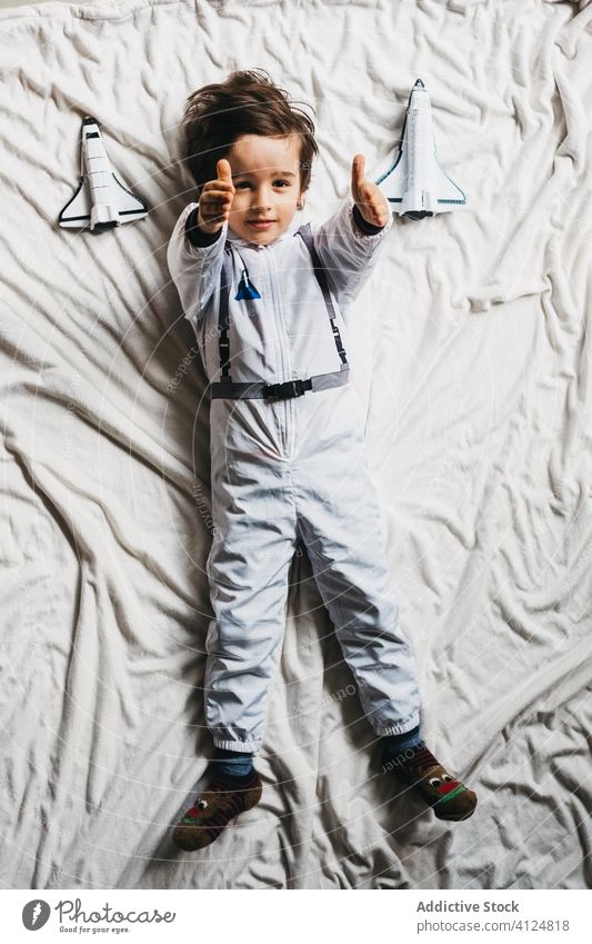 Boy in astronaut costume lying on bed boy spacesuit toy spaceship having fun smile kid cosmonaut bedroom child cheerful weekend rocket playful uniform pretend