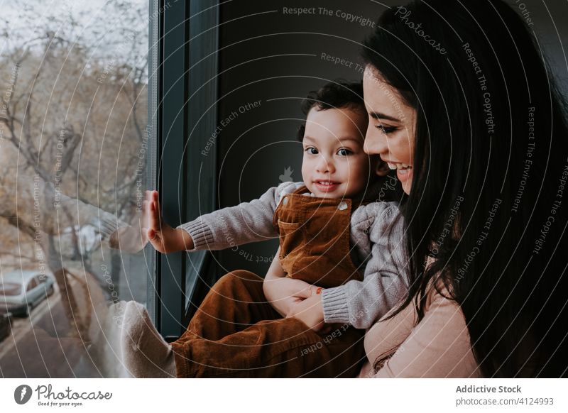 Positive young mother and little son standing near window and admiring view home curious explore point content excited cute child smile embrace kid parent