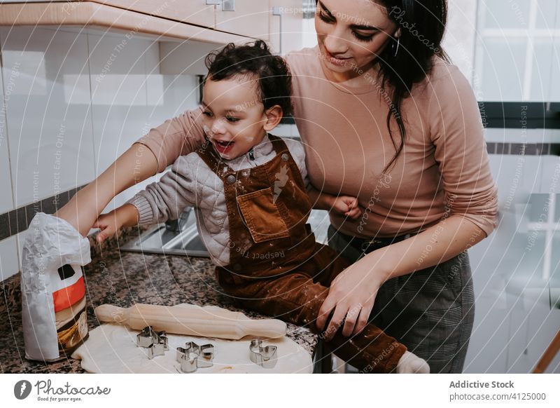 Cheerful mother cooking pastry with little son in kitchen rolling pin dough cookie helper assistant together bonding love chair kid cheerful flour woman food