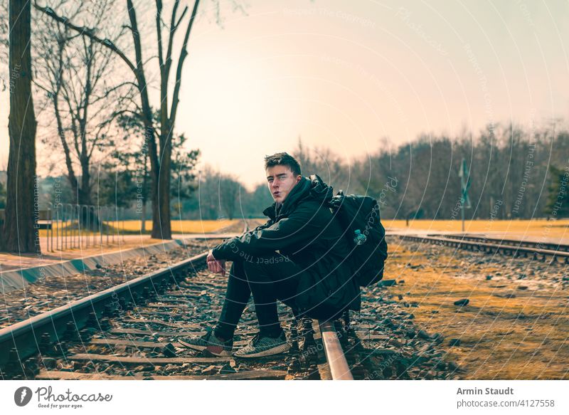 happy young man with backpack sitting on train tracks youthful Man Sit Railroad Tracks Backpack Landscape Outdoors fortunate Smiling Self-confident Powerful far