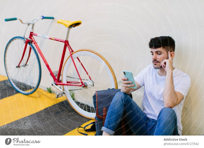 Man checking his smartphone sitting on the street floor bicycle bike fixie urban wheel fixed sport transportation gear lifestyle wall hipster ride pedal man