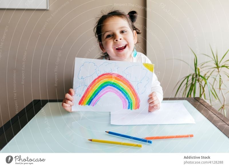 Cheerful girl showing drawing to camera cheerful colorful rainbow positive optimist charming table paper child smile cute hobby fun paint joy creative happy
