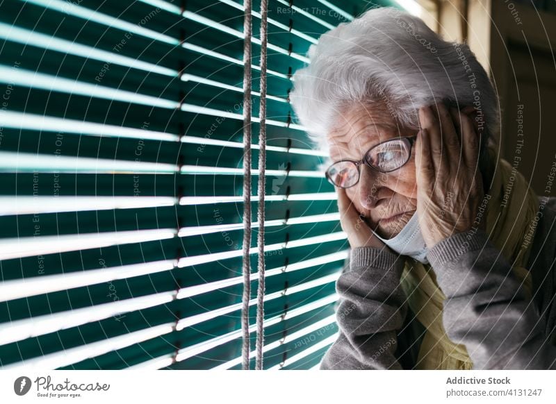 Anxious elderly woman standing near window during pandemic senior coronavirus stay home lonely unhappy sad shutter pensive quarantine mask prevent old female