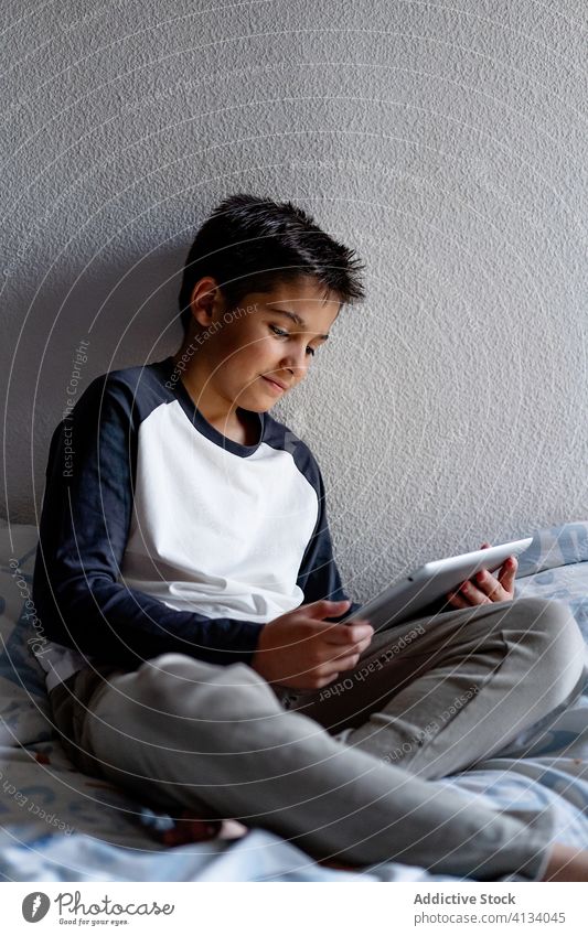 Delighted boy using tablet in bedroom during weekend kid blanket night watching cheerful pajama home adorable device gadget child internet browsing cozy rest