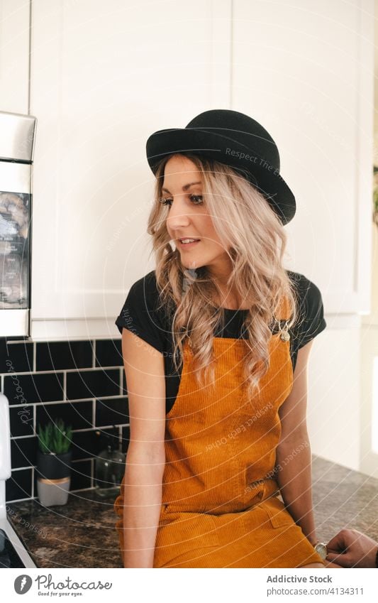 Thoughtful young woman in hat sitting on table in kitchen thoughtful counter hipster style modern alone think dreamy calm ponder pensive wistful contemplate