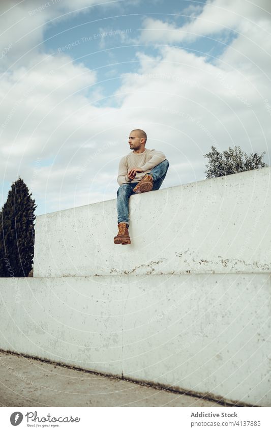 Hipster man sitting on concrete wall pensive hipster style trendy modern street style thoughtful contemplate fence young unshaven guy jeans casual sky cloudy