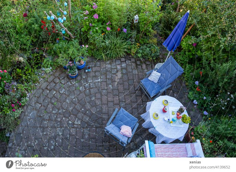 Bird's eye view in summer: Two yellow cups of coffee on the table, chairs with comfortable cushions and a parasol are ready on the terrace. This is surrounded by green plants with colorful flowers. Where have the coffee drinkers gone?