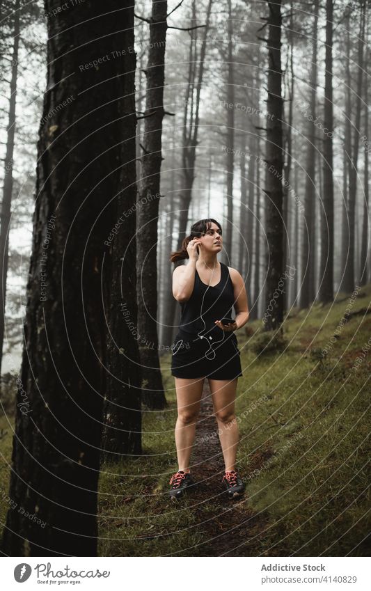Female runner listening to music in misty wood woman smartphone tired break using training forest female woods fog narrow activewear sportswoman path healthy
