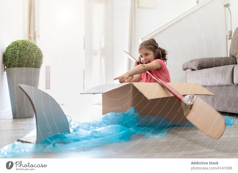 Joyful small girl in carton box sailing away from shark boat paddle game imagination fun playful kid sailor home child having fun joy pretend float childhood