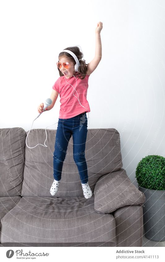 Happy preteen girl in headphones with mic singing while playing alone at home microphone having fun child jump sofa singer superstar happy music kid dance