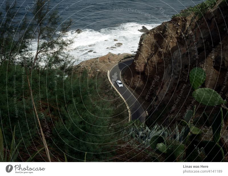 A great landscape of the northern Tenerife coastline. Rocky mountains, calm ocean, green plants, and remote roads. Somebody is traveling this tremendous path in a white car. Vacation postcard from an adventurous Tenerife.
