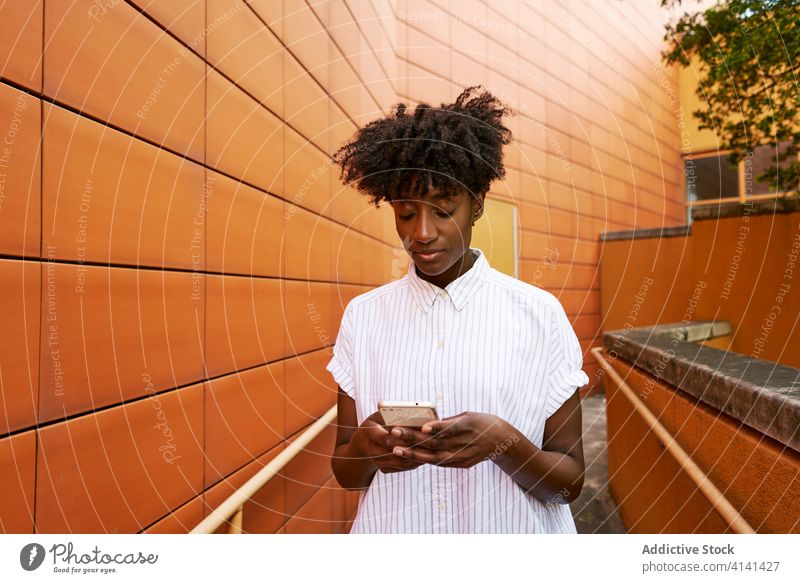 Focused black woman using smartphone on street against vivid exterior of modern building messaging pathway chill browsing smile social media facade surfing read