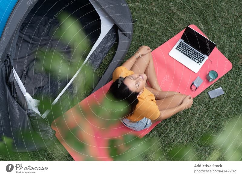 Happy woman with gadgets resting near tourist tent camp relax summer enjoy modern nature meditate meadow grass young female casual laptop device vacation travel