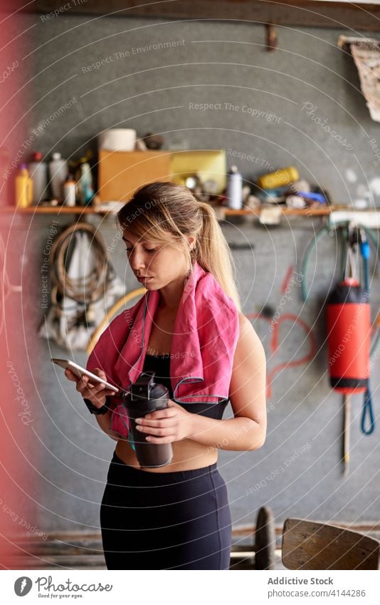 Focused sportswoman using smartphone in garage gym focus concentrate bottle break fit gadget browsing cellphone hydrate vitality training rest intense