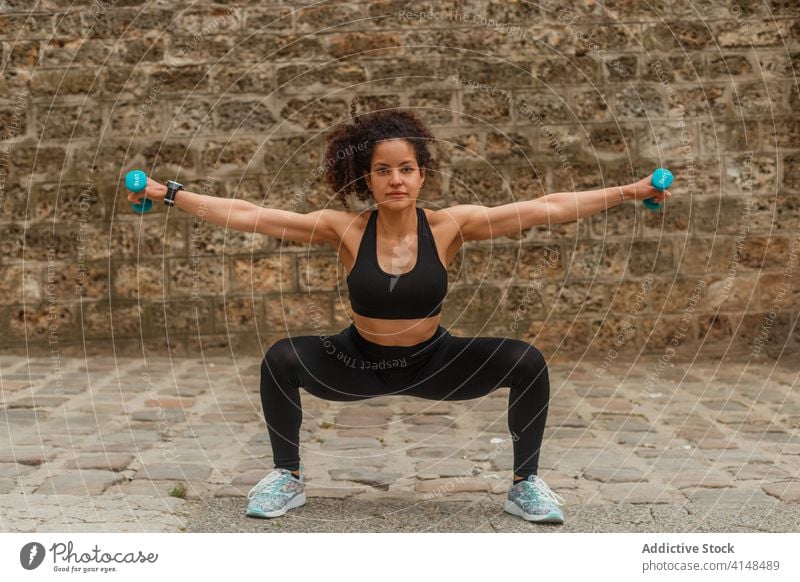 Fit ethnic sportswoman squatting with dumbbells on embankment during workout training balance reached arm pavement stretch practice flexible activewear strong