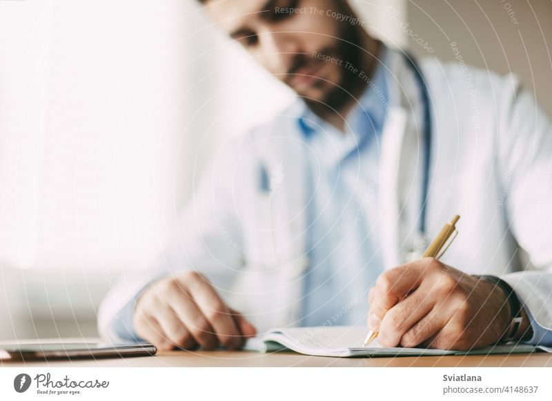 Friendly young male doctor of oriental appearance sits at a desk in his office and takes notes in a notebook, close-up medicine person health clinic stethoscope