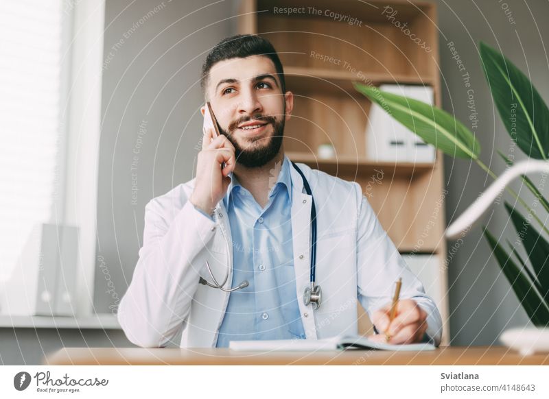 A young doctor of oriental appearance is talking on the phone, sitting at the table and making notes in a notebook medicine man health medical professional